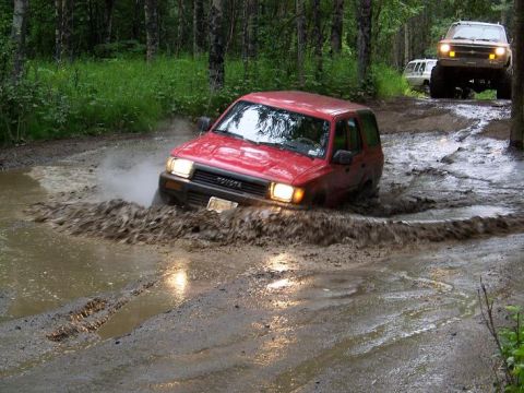 1985 Ford Bronco - A red Toyota SUV navigates a muddy trail, with other off-road vehicles following in the background.