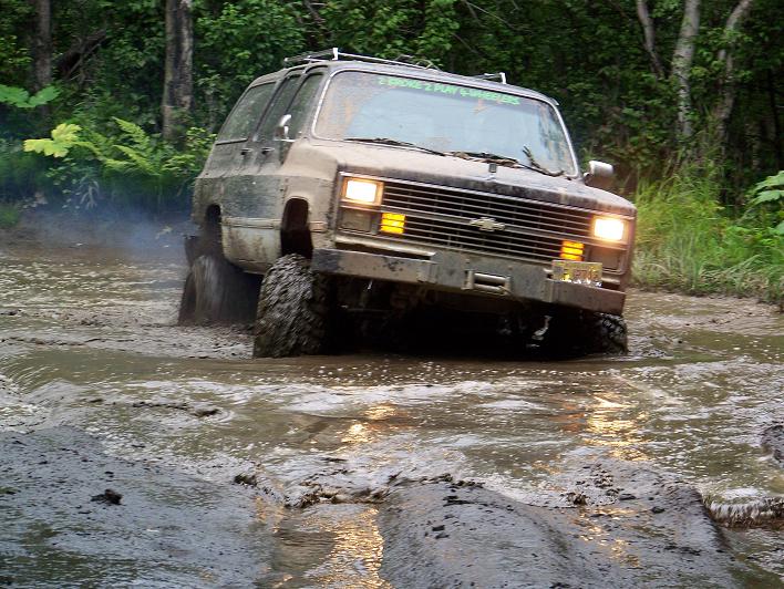 Muddy square-body Chevy Blazer driving through a flooded mud pit in the woods