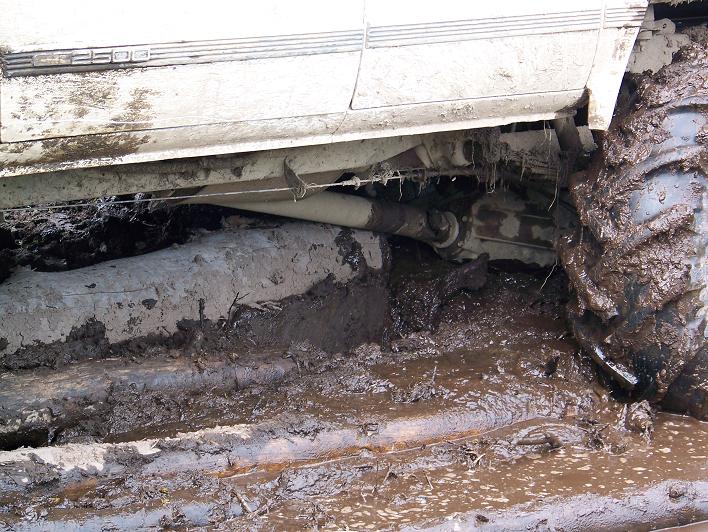 Underside of a mud-covered white truck with 2500 badge, driveshaft visible over a log-filled mud bog