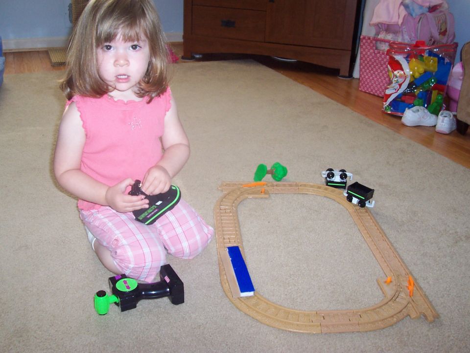 Child in pink shirt and plaid pants kneels on carpet with a toy train set