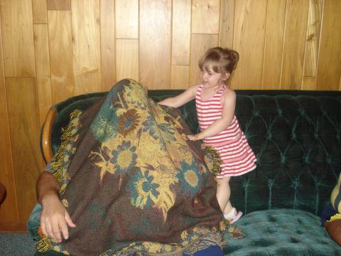 2000 Ford Crown Victoria - A young girl in a striped dress stands next to a person covered by a patterned blanket on a dark green tufted couch.