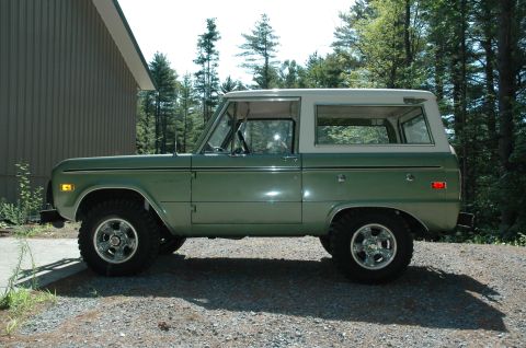 1973 Ford Bronco - A classic green Ford Bronco with a white top sits parked on a gravel surface, with trees and a building in the background.