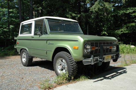 1973 Ford Bronco - A green 1973 Ford Bronco with a white top is parked on a gravel surface, with trees visible behind it.