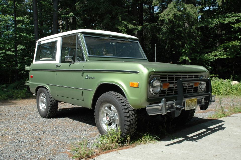Green early Ford Bronco with white hardtop and chrome winch bumper on gravel drive