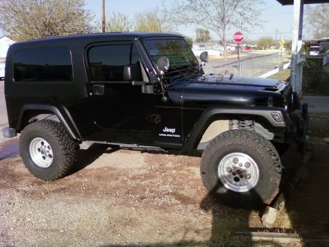 1985 Ford Bronco - This black Jeep Wrangler Unlimited is parked on a gravel surface under a clear sky.