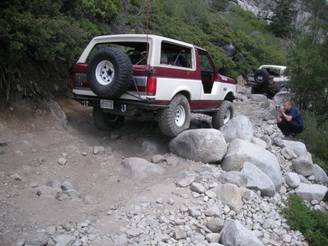 1991 Ford Bronco - A two-tone Ford Bronco tackles a challenging, rocky off-road path with another Bronco visible behind it.