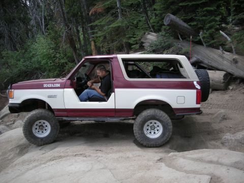 1991 Ford Bronco - A man drives a two-tone maroon and white Ford Bronco on a challenging off-road path.