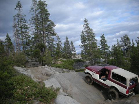 1991 Ford Bronco - A two-tone Ford Bronco navigates a challenging off-road course amidst a rocky, wooded landscape.