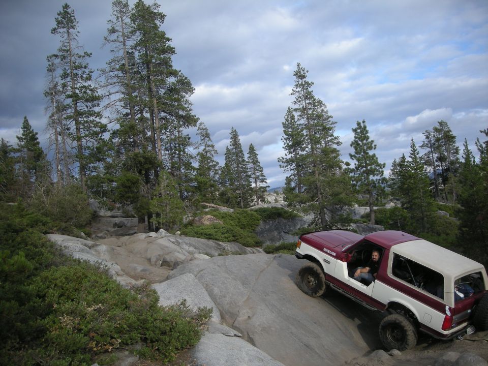 Ford Bronco climbing a steep, rocky trail with trees and cloudy sky in the background