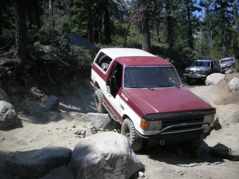 1991 Ford Bronco - This red and white SUV navigates a challenging rocky trail, with other vehicles following behind.