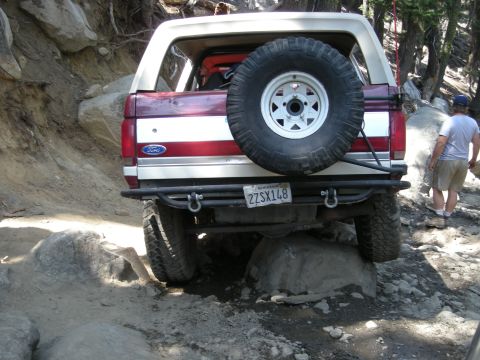1991 Ford Bronco - 1991 Ford Bronco navigating rocky terrain, rear view shows spare tire and custom bumper.