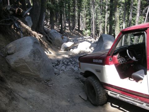 1991 Ford Bronco - A two-tone truck navigates a challenging, rocky path through a dense forest.
