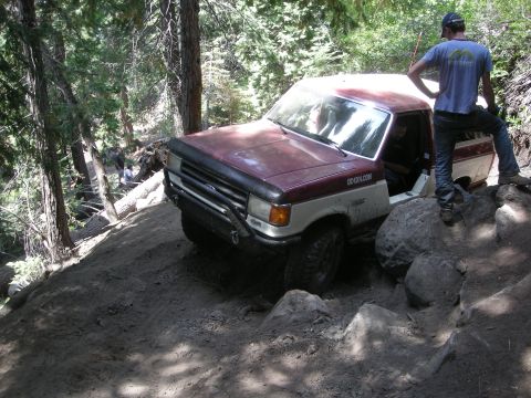 1991 Ford Bronco - This two-tone pickup truck is navigating a challenging off-road trail with large rocks and steep inclines.