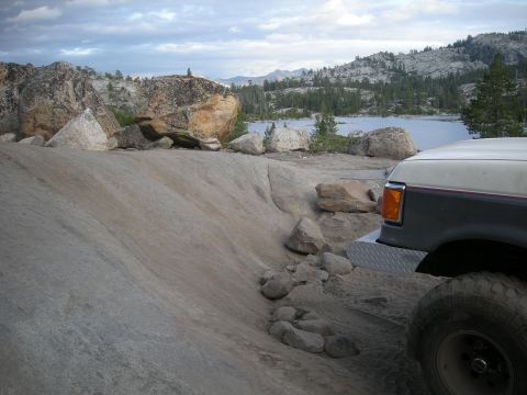 1991 Ford Bronco - The front corner of a 1991 Ford Bronco is visible, parked on a rocky terrain with a lake and mountains in the background.