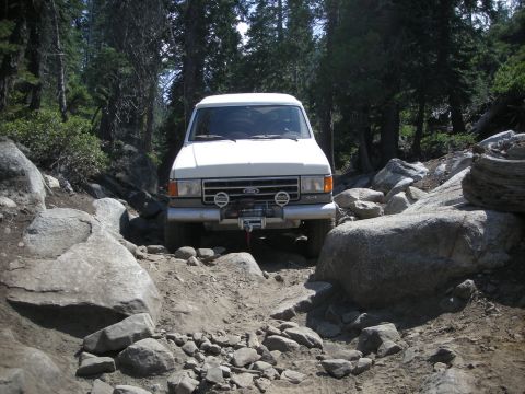1991 Ford Bronco - A white Ford Bronco is shown on a challenging off-road trail surrounded by trees and large rocks.