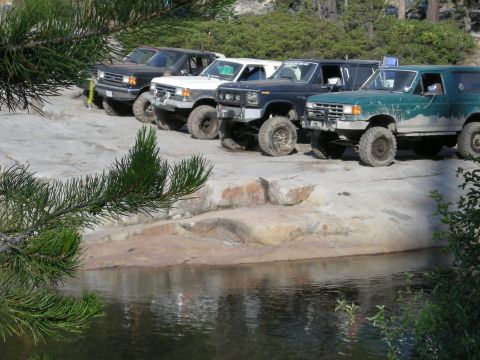 1991 Ford Bronco - Several lifted Ford Broncos are parked in a rocky area next to a body of water.