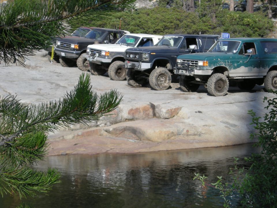 Four modified Ford Broncos parked on rocks near water, one green, one black, one white, one dark