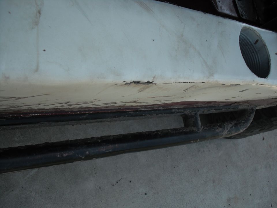 Close-up of a white vehicle bumper with a black rock slider underneath. A circular opening is visible on the right side of the bumper