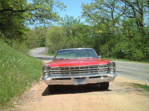 1967 Ford Galaxie - A red convertible sits by the side of a road on a sunny day, with trees and greenery surrounding it.