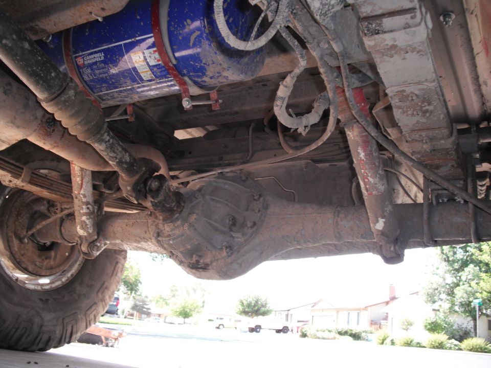 Undercarriage view showing a large blue tank, driveshaft, differential, and leaf springs. Text is visible on the blue tank