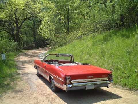 1967 Ford Galaxie - A red 1967 Ford Galaxie convertible is parked on a dirt road surrounded by trees and grass.