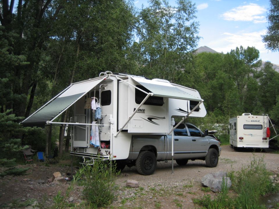 Silver dually pickup with white Lance truck camper, awnings out, at a forested campsite