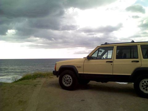 1985 Ford Bronco - A tan Jeep Cherokee XJ is parked on a dirt path overlooking the ocean under a cloudy sky.