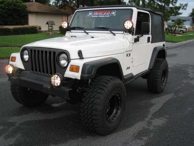 White Jeep Wrangler TJ with aftermarket bumper, lights, and off-road tires. Decal on windshield reads 'TEAM 4X4'
