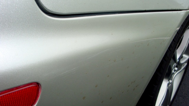 Silver car quarter panel with numerous scratches and brown spots, showing a red taillight and part of a wheel