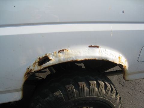 1996 Ford Bronco - Heavily rusted rear wheel well of a white SUV shows significant metal decay and a portion of the tire.