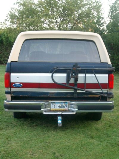 1987 Ford Bronco - The rear of a 1987 Ford Bronco is shown with a blue and red striped tailgate and a tan removable roof.