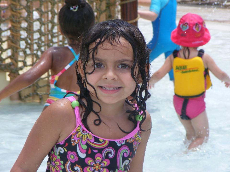 Girl in floral swimsuit smiles in shallow water, other kids in background