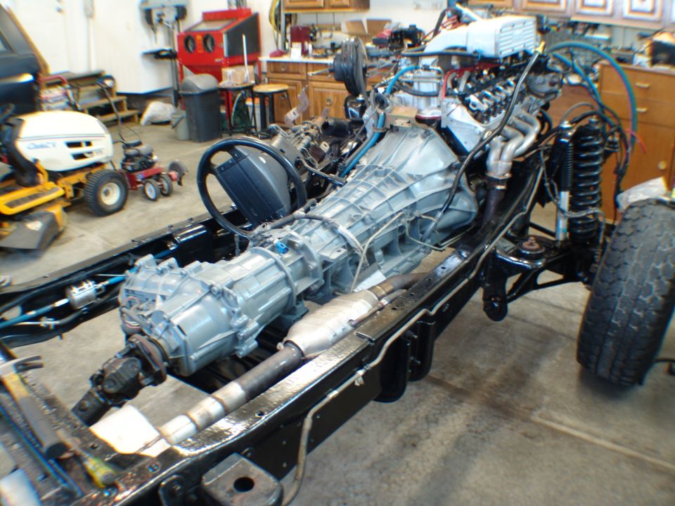 Vehicle frame with engine, transmission, steering wheel, and suspension components visible. A lawnmower is in the background