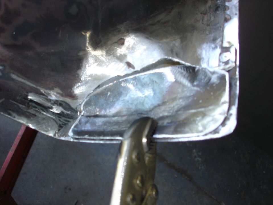 Metal panel with visible welding seams and a clamp holding a section. The panel is shiny and appears to be undergoing repair