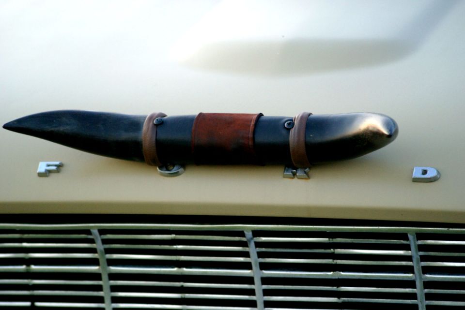 Tan car hood with a black, horn-shaped hood ornament secured by leather straps, and chrome 'F' and 'D' emblems