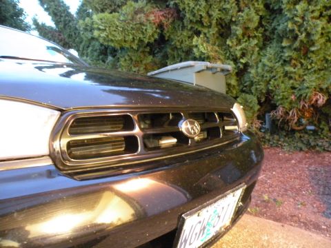 1998 Subaru Legacy - Close-up of a car's front grille with a Subaru emblem, parked outdoors near trees.
