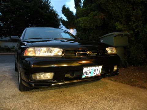 1998 Subaru Legacy - The front grille and headlights of a black Subaru sedan are visible, parked near a trash bin.