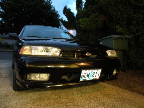 1998 Subaru Legacy - A black 1998 Subaru Legacy sedan is parked in front of trees and a trash bin.