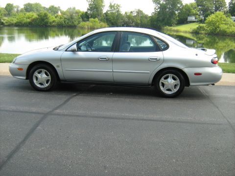 1988 Mercury Grand Marquis - This silver sedan is parked on a road next to a calm lake with green trees on the opposite bank.