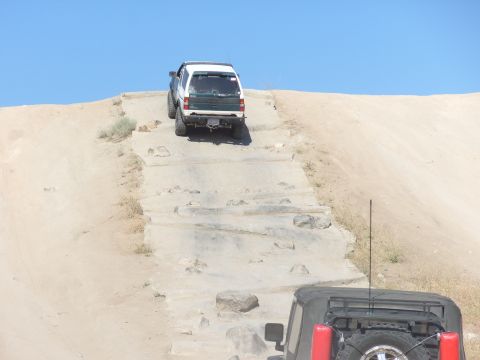 1995 Ford Bronco - Two SUVs navigate a challenging off-road course with a steep, rocky incline under a clear blue sky.
