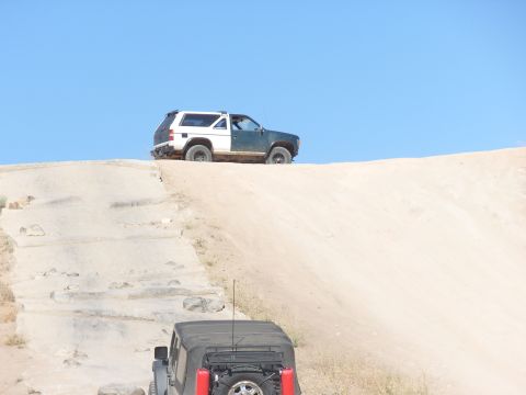 1995 Ford Bronco - Two off-road vehicles ascend a steep, sandy incline under a clear blue sky.