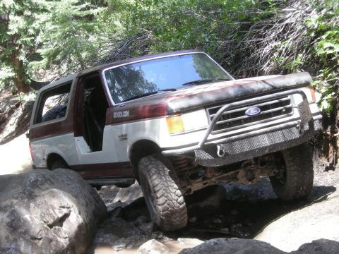1991 Ford Bronco - This modified Ford Bronco is tackling a challenging off-road path with large rocks and trees.