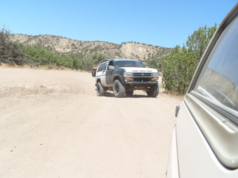 1995 Ford Bronco - Two off-road vehicles navigate a dusty trail in a dry, hilly landscape under a clear blue sky.