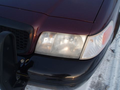 1999 Ford Crown Victoria - Front corner of a dark red car with a black grille and bumper, showing the headlight assembly.