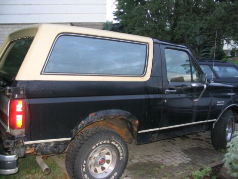 1986 Ford Bronco - Side view of a black Ford Bronco with a tan removable top, parked outdoors.