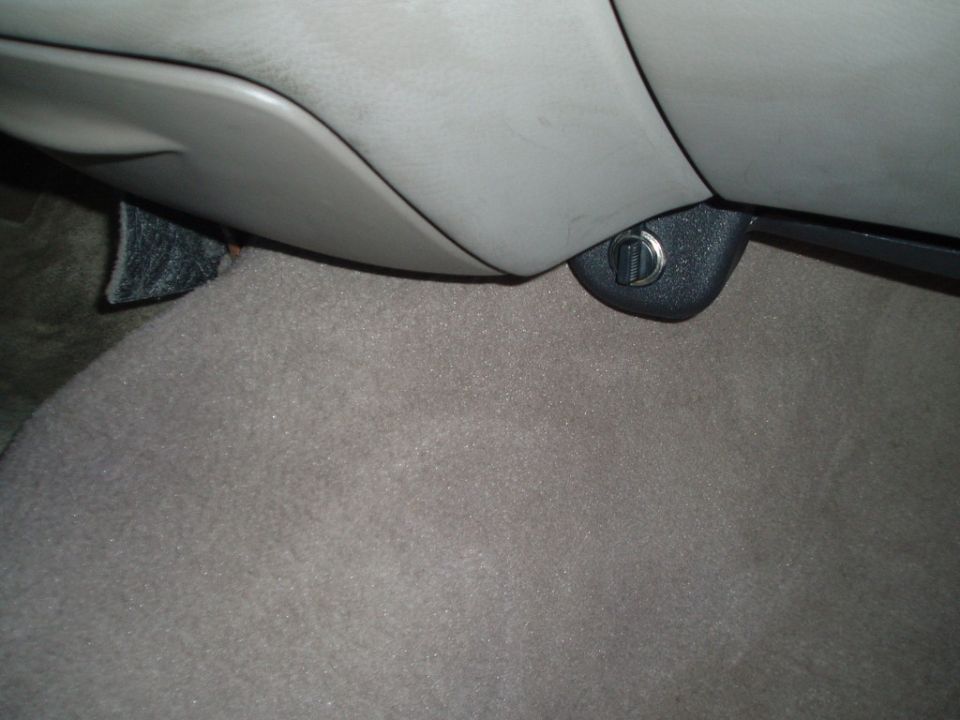 Car interior with beige carpet and gray plastic trim, showing a black latch with a silver D-ring