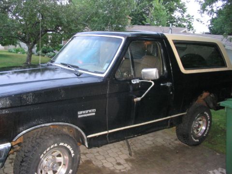 1986 Ford Bronco - This black Ford Bronco with a tan top is viewed from the side, revealing its chrome trim and a slightly worn appearance.