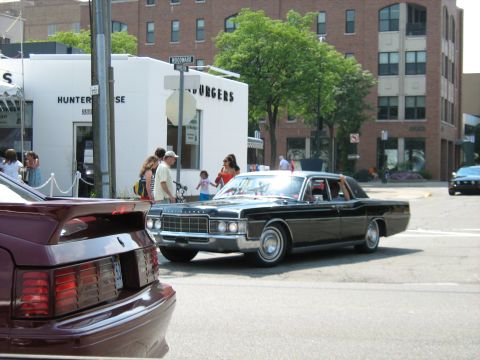 1988 Mercury Grand Marquis - A black sedan drives past a building with "BURGERS" signage. Another car is visible in the foreground.