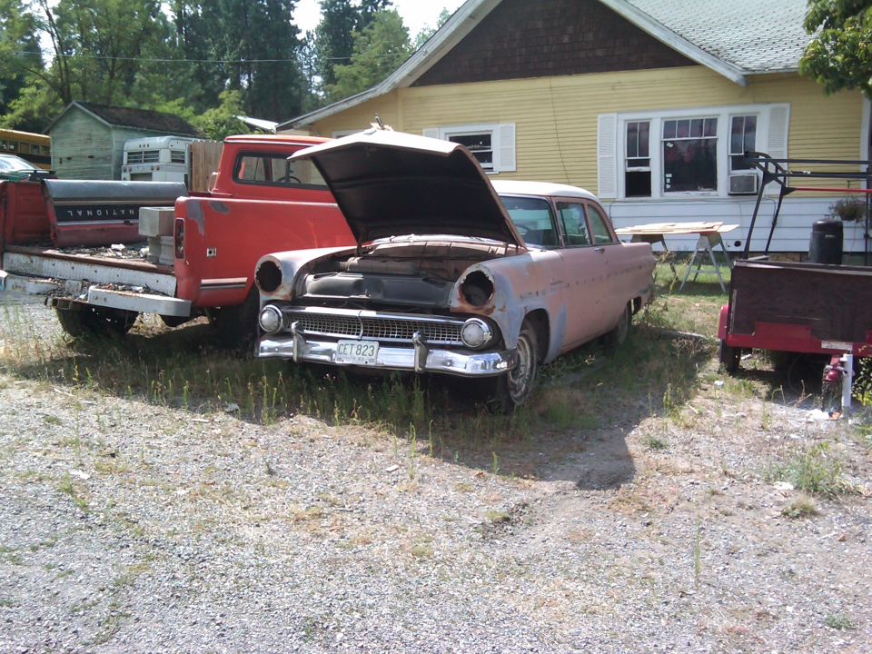 Rusty sedan with hood open, missing headlights, parked next to a red truck