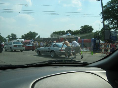 1988 Mercury Grand Marquis - Two men push a silver coupe down the street during an event, with tents and spectators visible.
